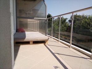 A balcony with a mattress and a red pillow, a glass railing, and a view of greenery. - Silver Beach B16 Apartment (Byala)