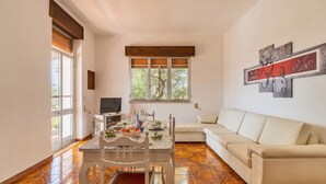 A spacious living room with a white sofa, a dining table set with glassware, and a large window with a view of trees. - 2929 Villa Garden Rose by Barbarhouse (Porto Cesareo)