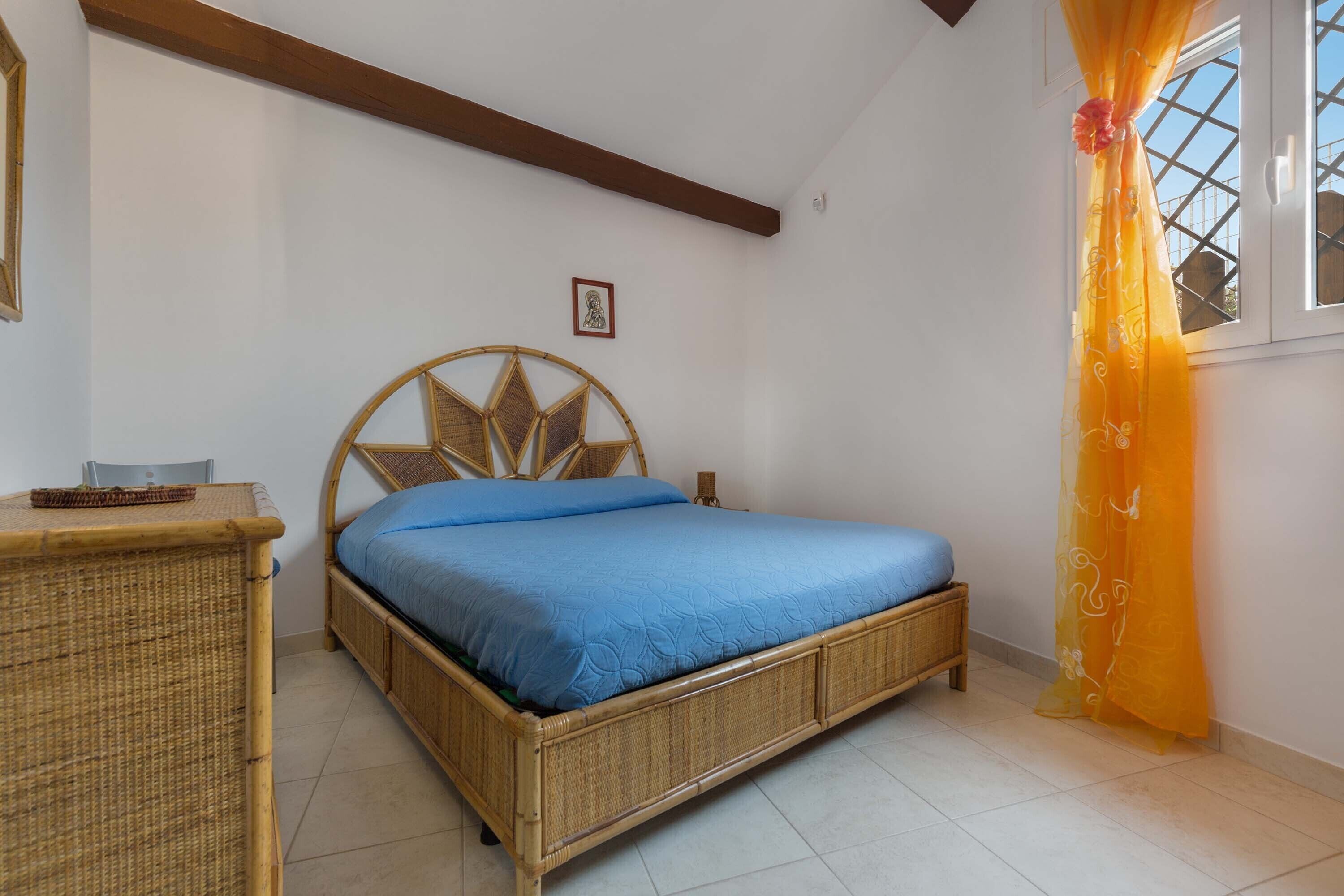 A room with a bed, a wicker dresser, and a window with a curtain.