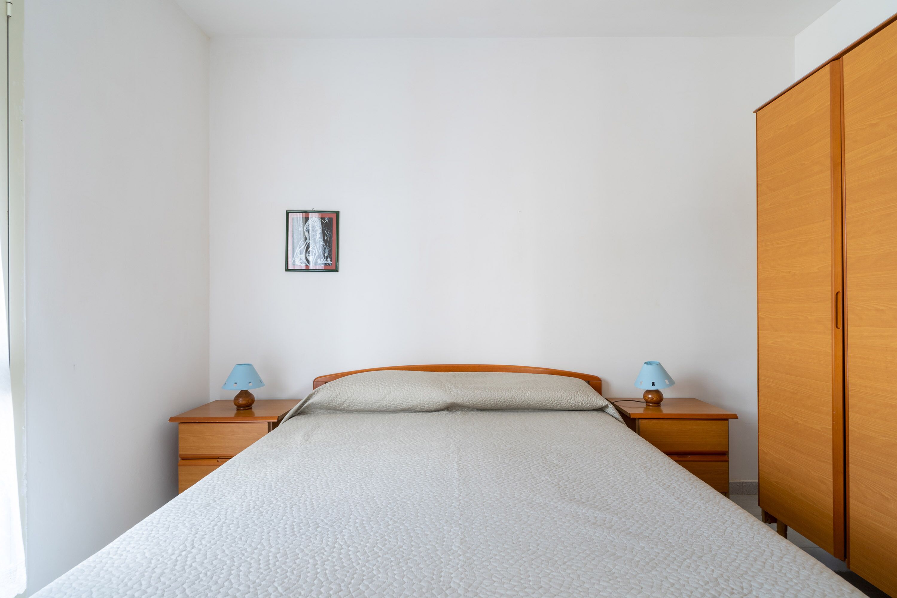 Un dormitorio con cama, dos mesitas de noche, un armario, un cuadro enmarcado en la pared y una ventana.