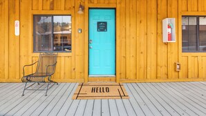 A wooden building with a turquoise door, a metal chair, and a 'HELLO' mat.