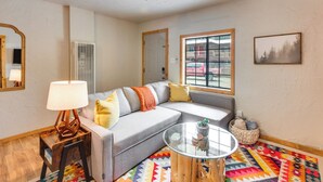 A living room with a gray sofa, a glass coffee table, a wooden side table with a lamp, a colorful rug, and a window with a view of a red car and buildings.