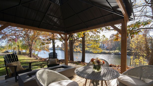 A covered outdoor seating area with wicker furniture, a table with a vase of flowers, and a view of a lake and autumn trees.