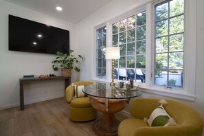 Un salon avec une table basse en verre, deux fauteuils jaunes, une table d’appoint en bois sur laquelle est posé un jeu d’échecs, une plante en pot et un téléviseur fixé au mur.