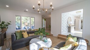 A spacious living room with a grey sofa, a round coffee table, and a chandelier. There is a dining area with a table and chairs, and a staircase leading to another level.