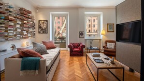 A modern living room with a sofa, armchair, and coffee table. There is a bookshelf filled with books, a television, and a window with a view of buildings. - Wonderful 1 Bedroom Apartament in the City Center (Rome)