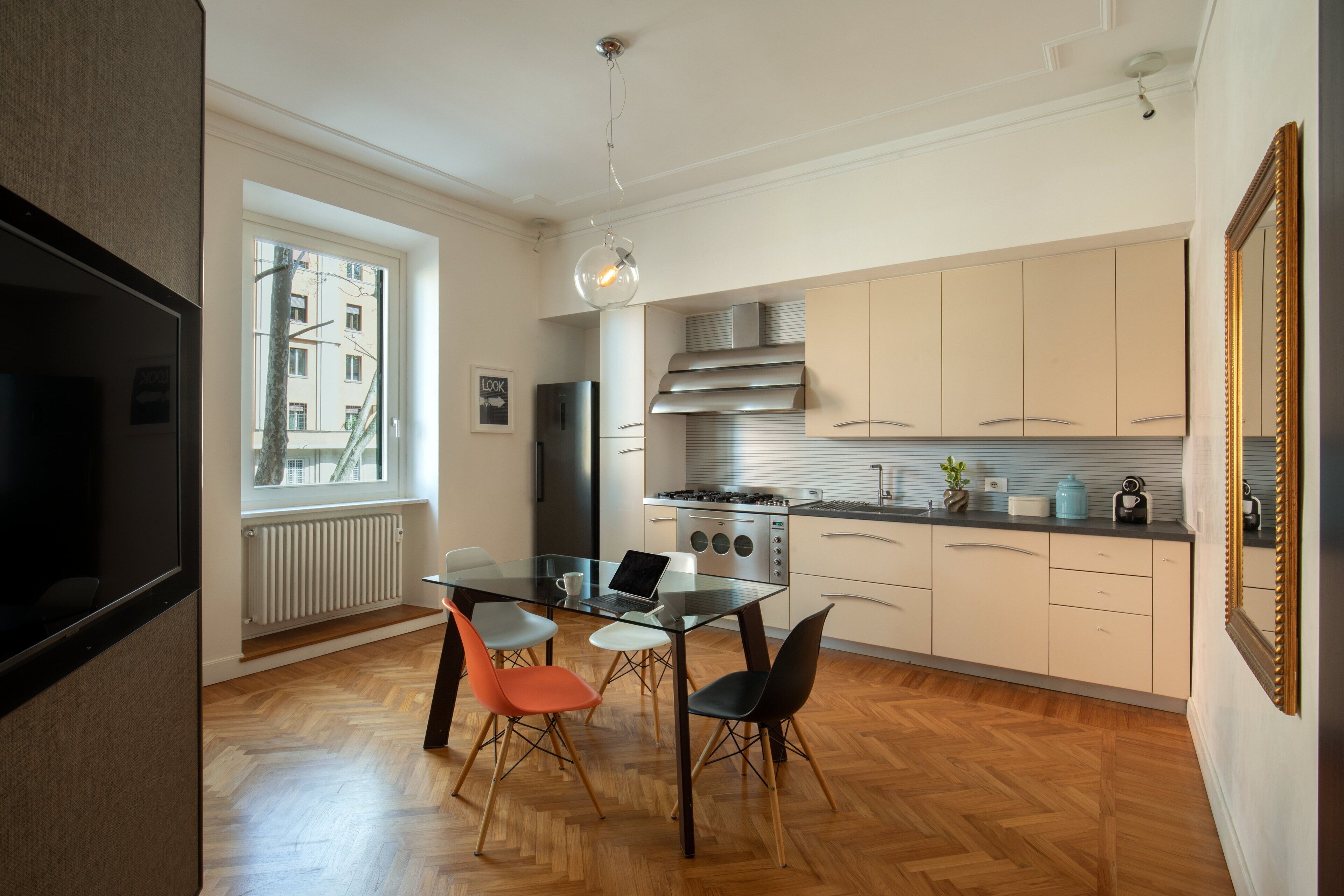 A modern kitchen with a dining area, featuring a glass table, wooden flooring, and a view of the city through a large window.