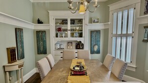 Comedor con mesa de madera, sillas tapizadas en tono beige, lámpara de araña y aparador empotrado con objetos decorativos.