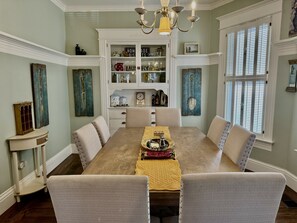 Una sala da pranzo con un tavolo in legno, sedie rivestite in color beige, un lampadario e un mobile incassato con oggetti decorativi.