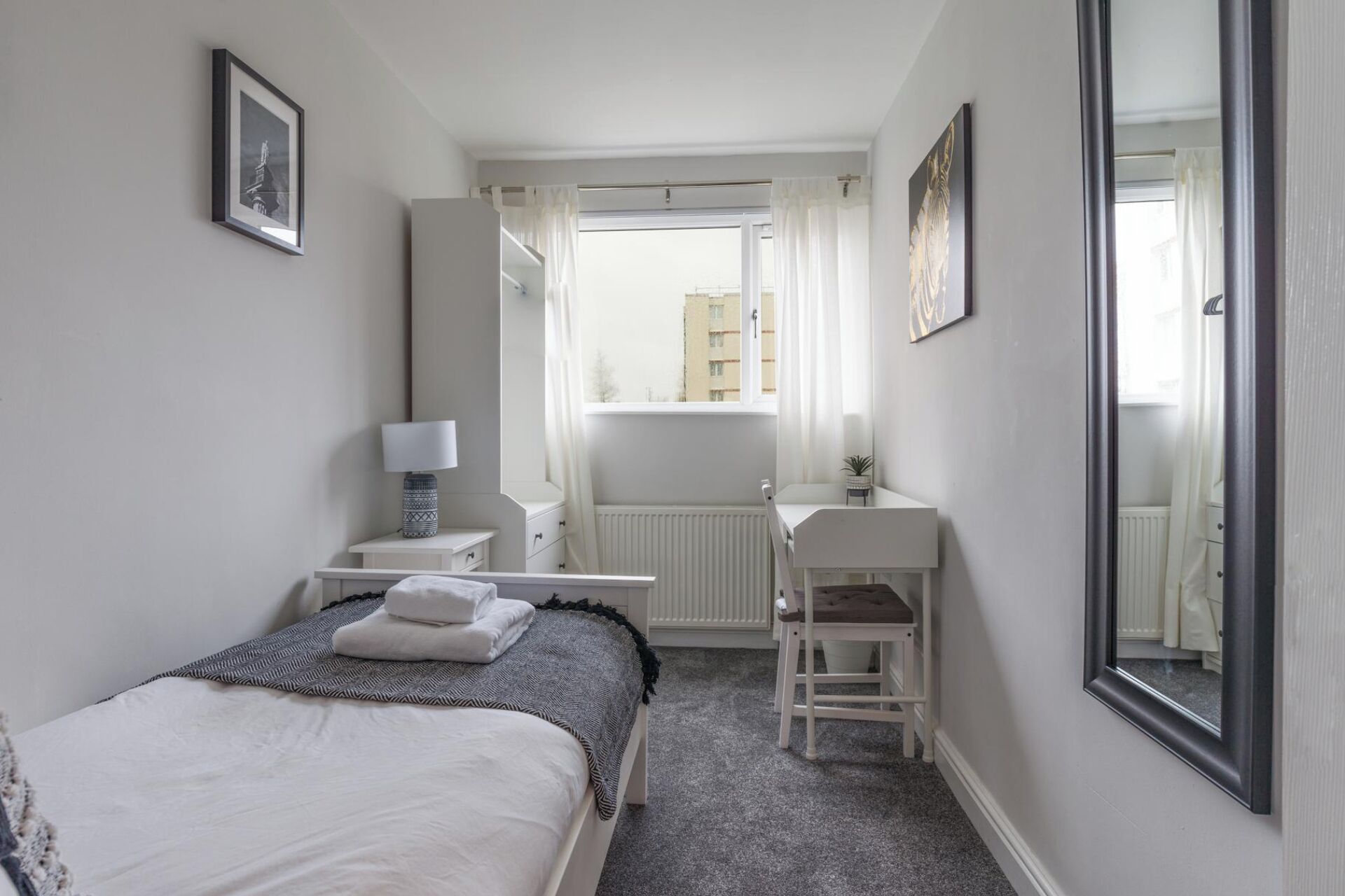Ein Hotelzimmer mit Bett, Schreibtisch, Stuhl, einem Fenster mit Vorhängen und einem großen Spiegel.