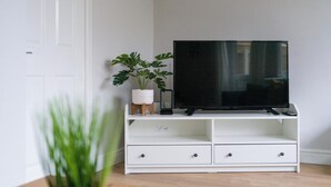 A white entertainment center with a flat-screen TV, a potted plant, and a small wooden side table. - Howick House-sleeps 5-pet Friendly-parking-garden (Newcastle-upon-Tyne)