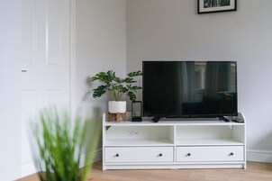 Un meuble de rangement blanc pour appareils électroniques, avec un téléviseur à écran plat, une plante en pot et une petite table d’appoint en bois.