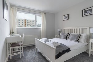 Une chambre à coucher avec un lit, des tables de chevet, une chaise, une fenêtre avec des rideaux et des tableaux encadrés accrochés au mur.