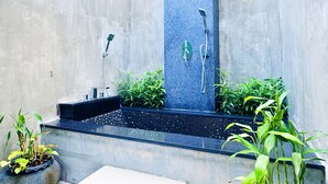 Un área de ducha exterior moderna con una fuente vertical de agua, un banco y plantas en macetas.