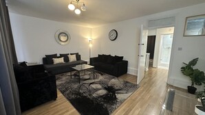 A modern living room with a black sofa, a glass coffee table, and a patterned rug. There are two wall clocks and a ceiling light fixture. - Gorgeous 4 Bedrooms, 5-baths House Central London (London)