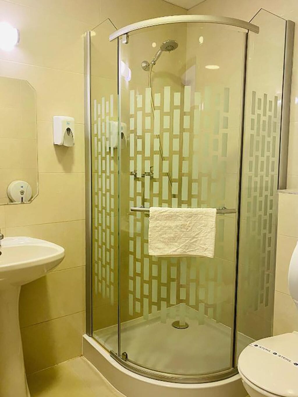Une cabine de douche fermée par des parois en verre, avec une serviette accrochée à la porte, un lavabo et des toilettes, le tout dans une salle de bain aux murs de couleur beige et dotée d’une porte en verre décorée.
