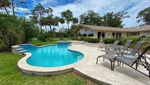 Una piscina de diseño curvo, rodeada por un patio con sillones de descanso y una casa al fondo.