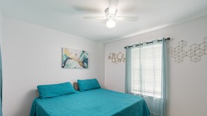 Een slaapkamer met een bed, een plafondventilator, een raam met gordijnen en wanddecoratie.