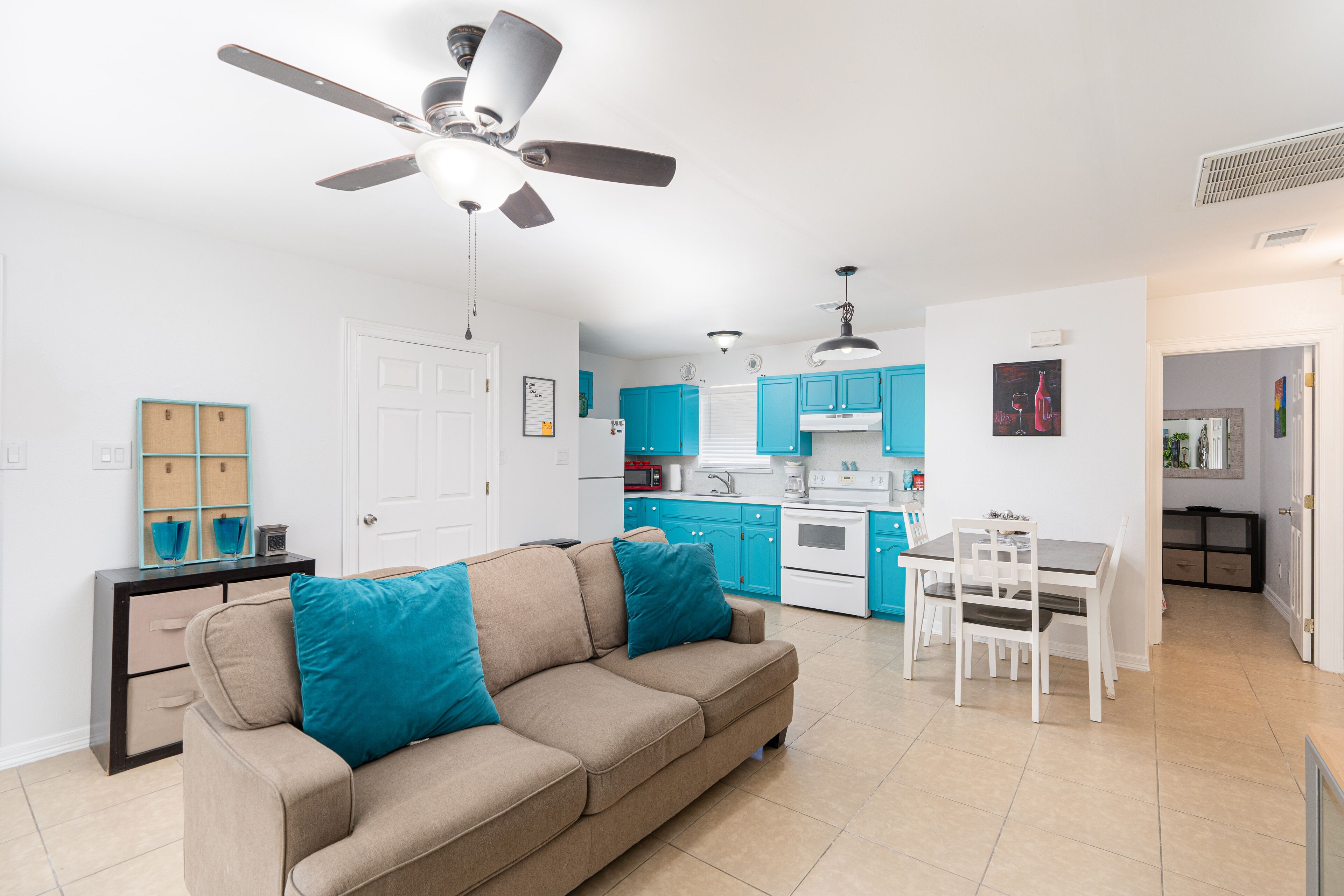 A modern living room with a beige sofa, teal cushions, a dining area with a white table and chairs, a kitchen with blue cabinets, and a ceiling fan.