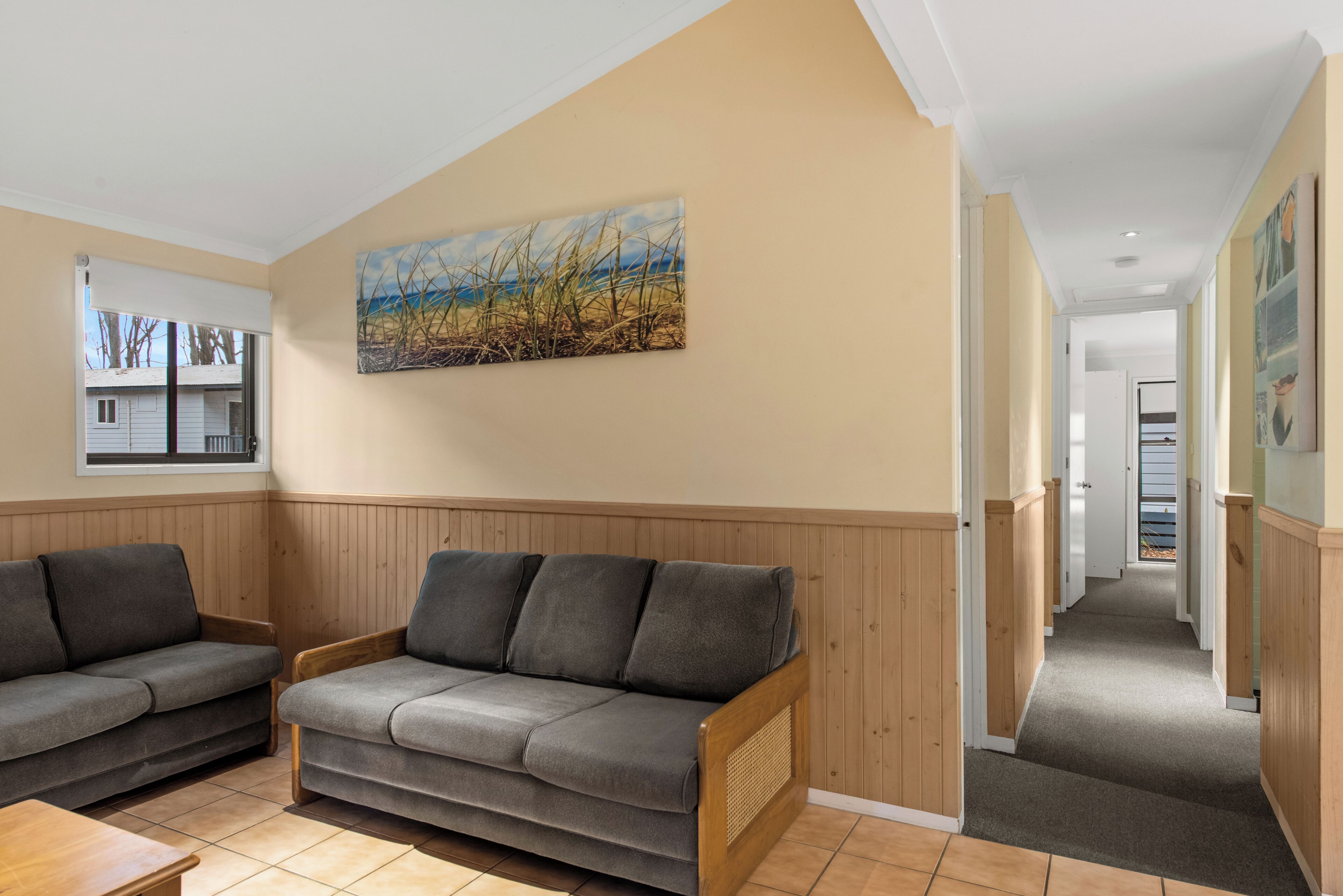 Une pièce avec deux canapés gris, un banc en bois, un tableau représentant un champ, et un couloir menant à une autre pièce.