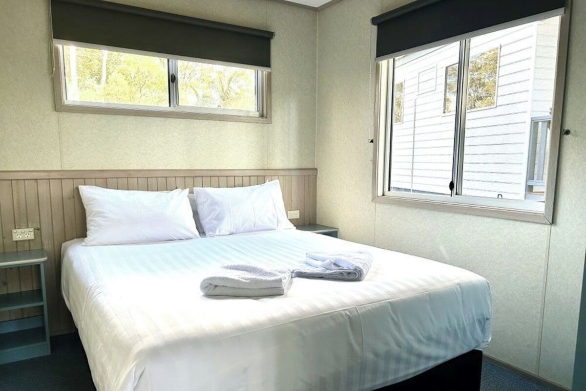 A neatly made bed with white linens and a folded towel, a bedside table, and a window with blinds.