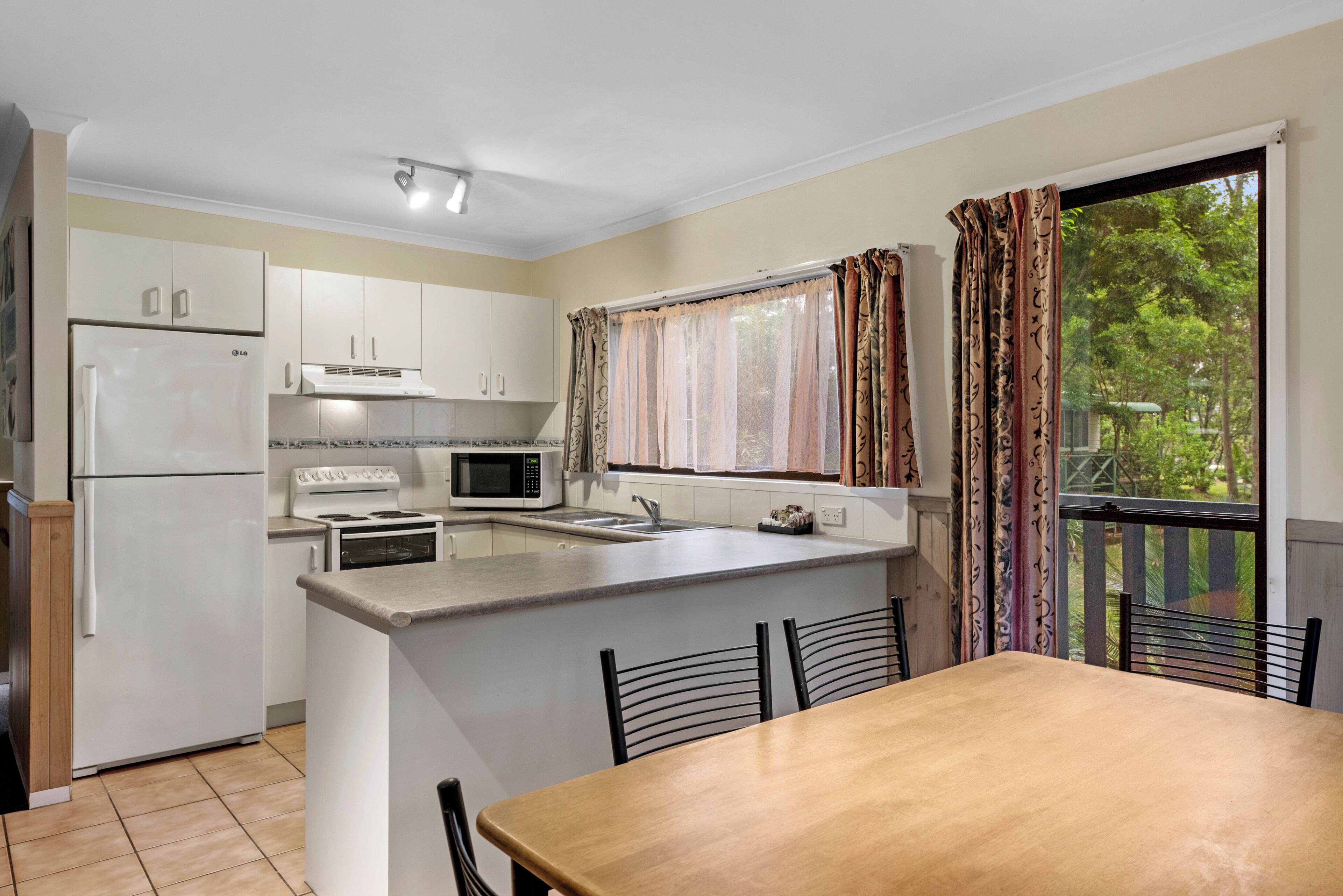 A compact kitchen with white cabinets, a refrigerator, a microwave, and a stove. There is a dining table with chairs and a window with curtains.