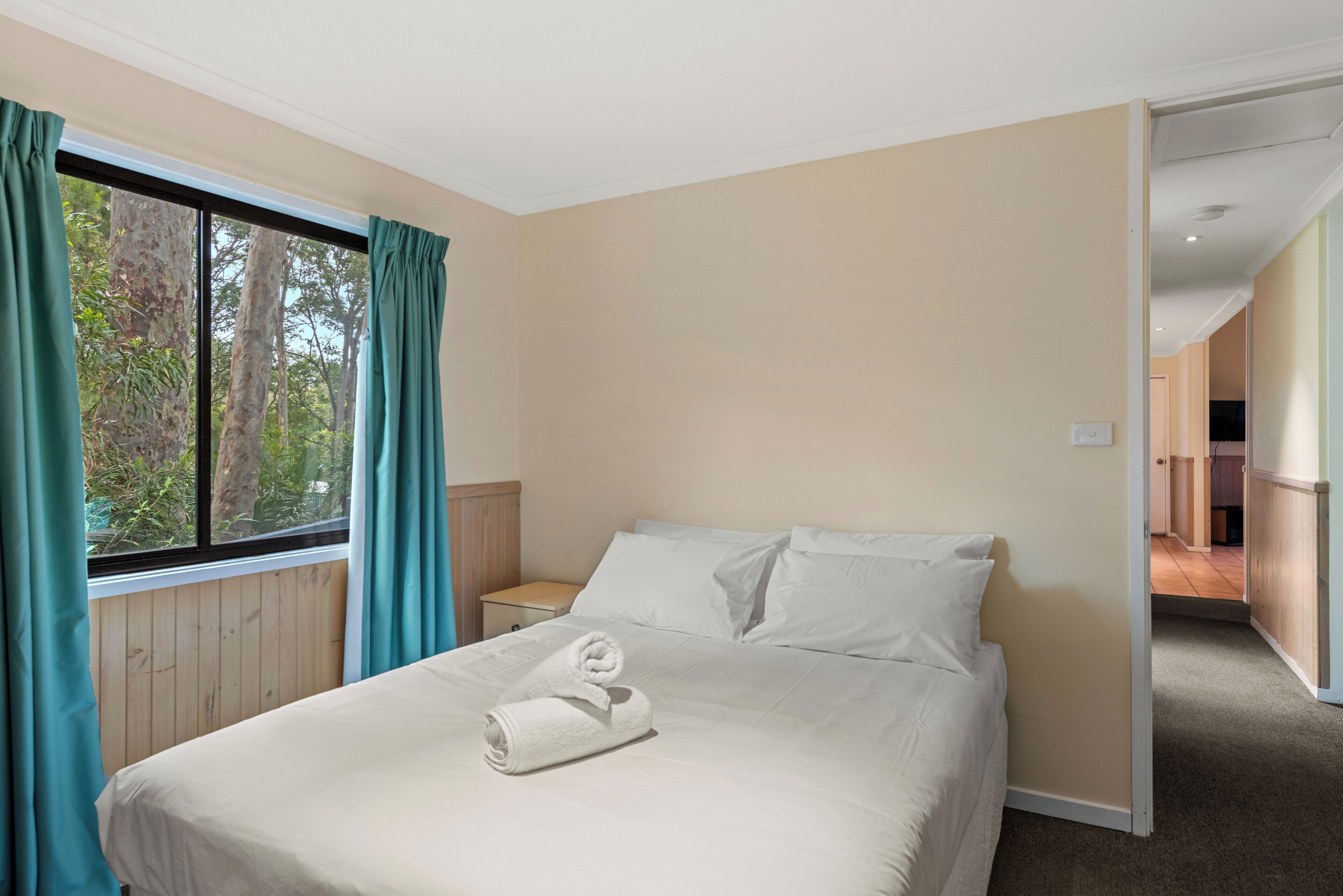 Habitación de hotel con cama, mesita de noche, ventana con cortinas y vistas a árboles.