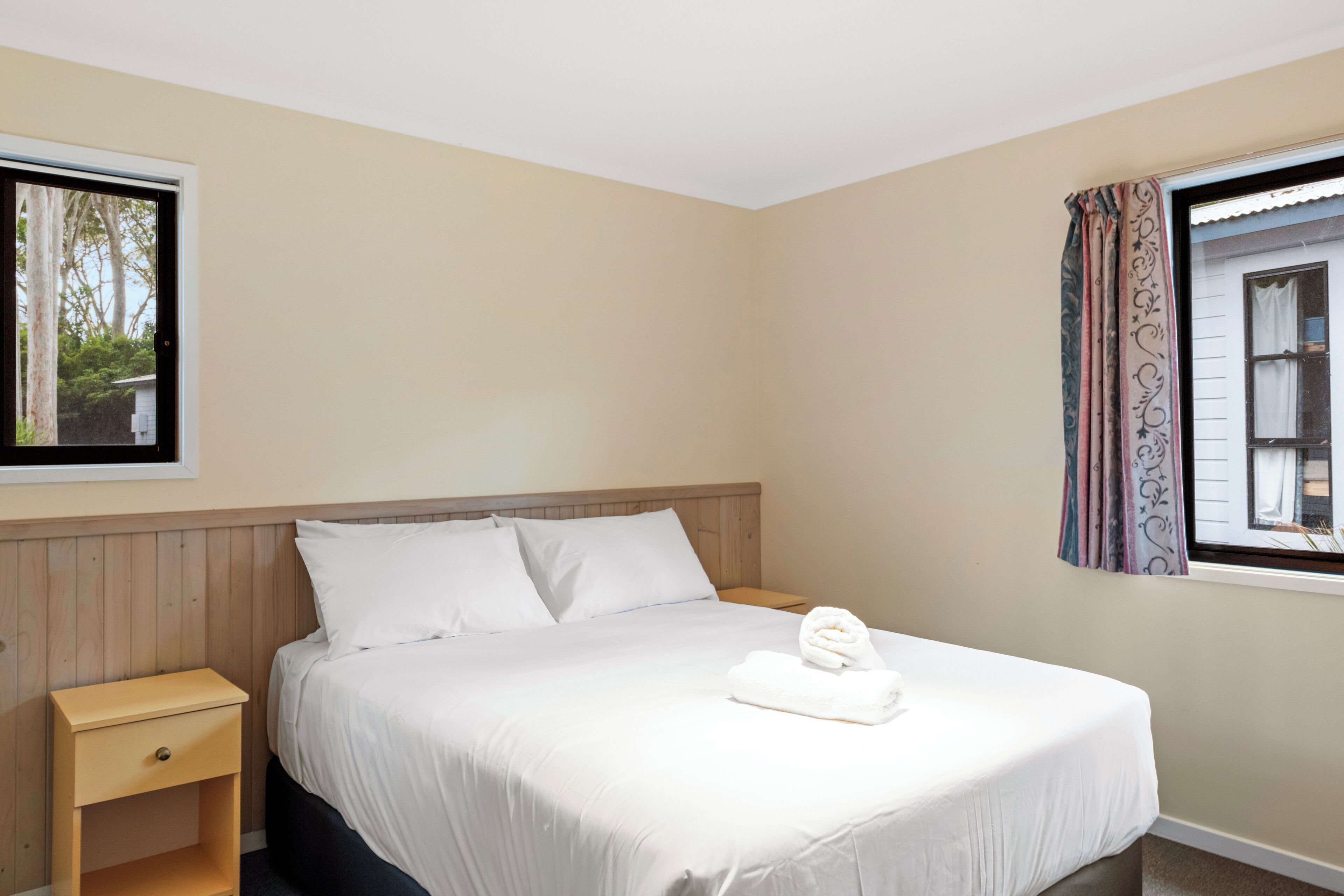 A neatly made bed with white linens and a rolled towel, a wooden nightstand with a drawer, a window with a view of trees, and a curtain with a floral pattern.