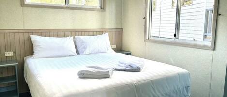 Una cama bien hecha con ropa de cama blanca y una toalla doblada, una mesita de noche y una ventana con persianas.