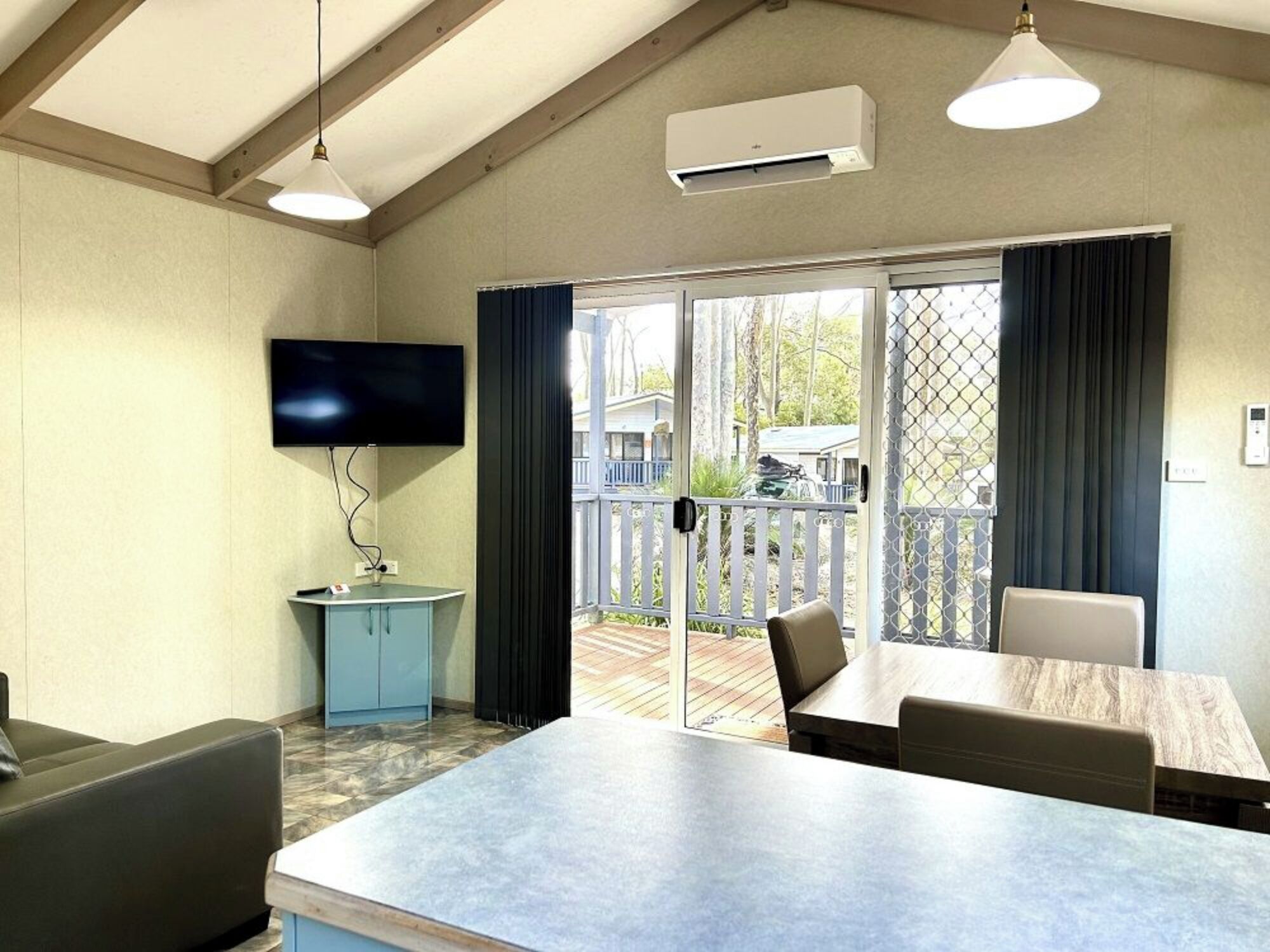 Een kamer met een flatscreen televisie aan de muur, een blauwe kast, een houten tafel met stoelen en een schuifdeur naar een terras.