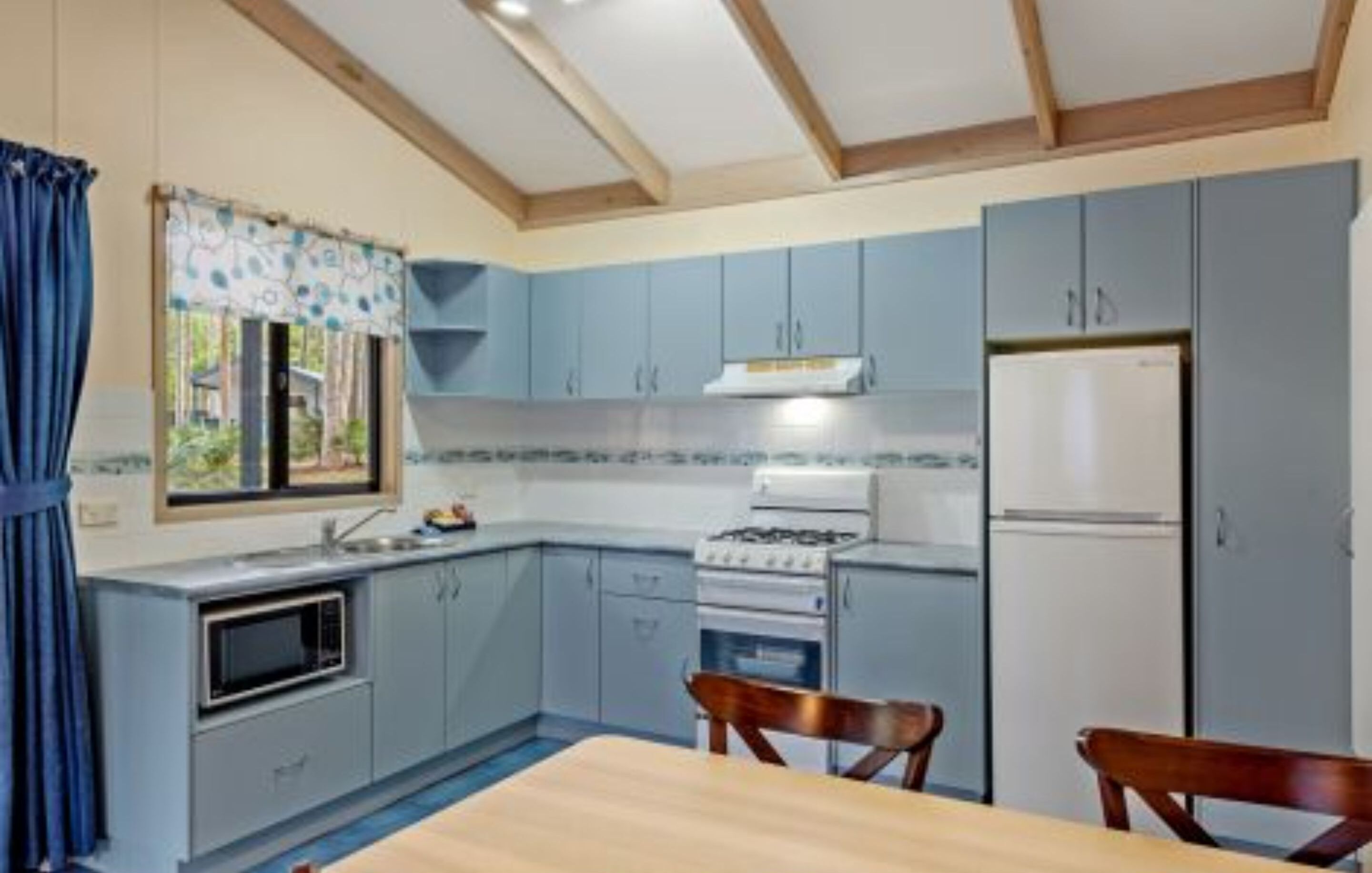 A compact kitchen with a gas stove, microwave, and refrigerator, wooden chairs, and a window with curtains.