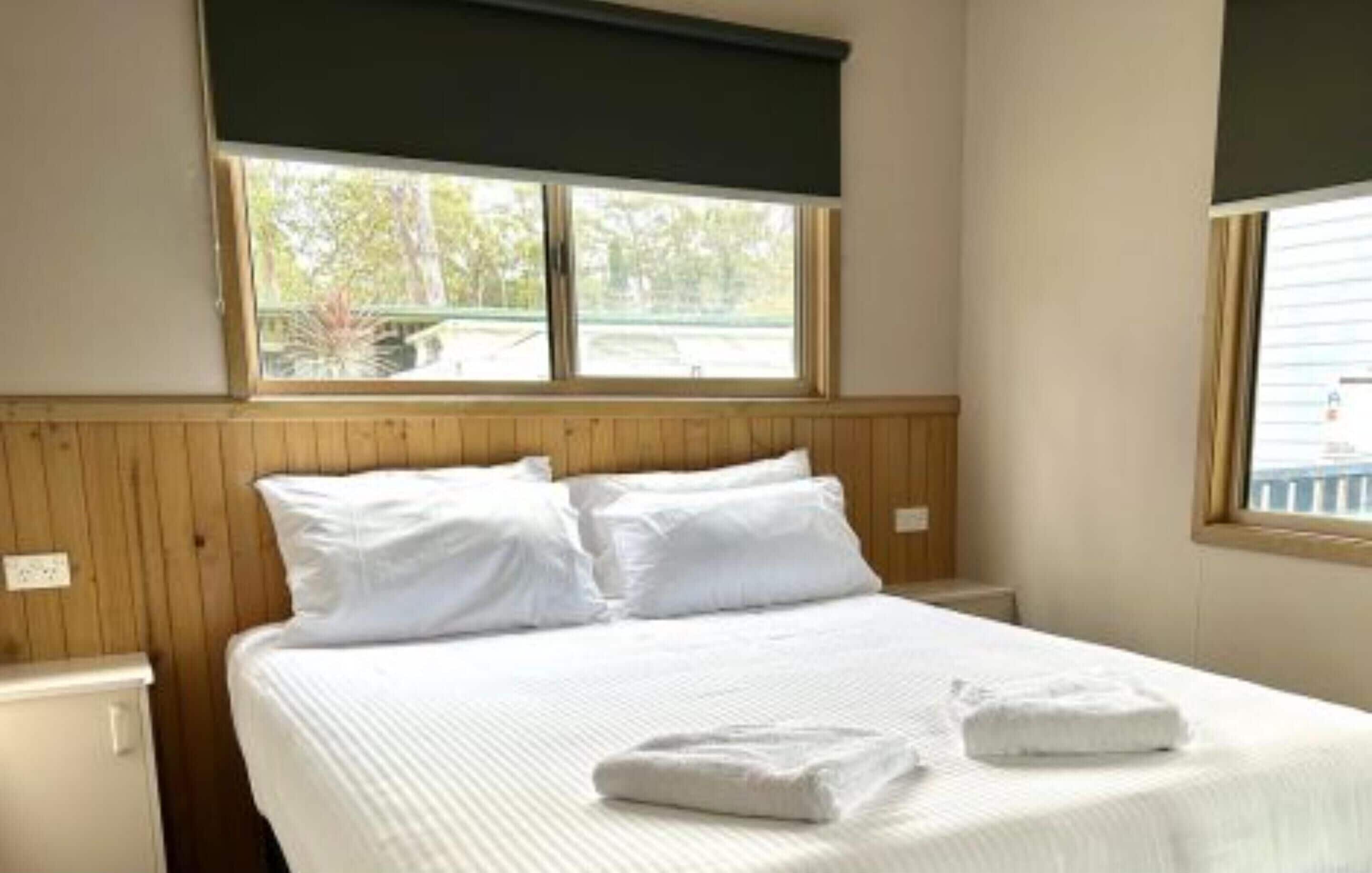 A neatly made bed with white linens and pillows, a window with a view of trees, and wooden paneling on the walls.