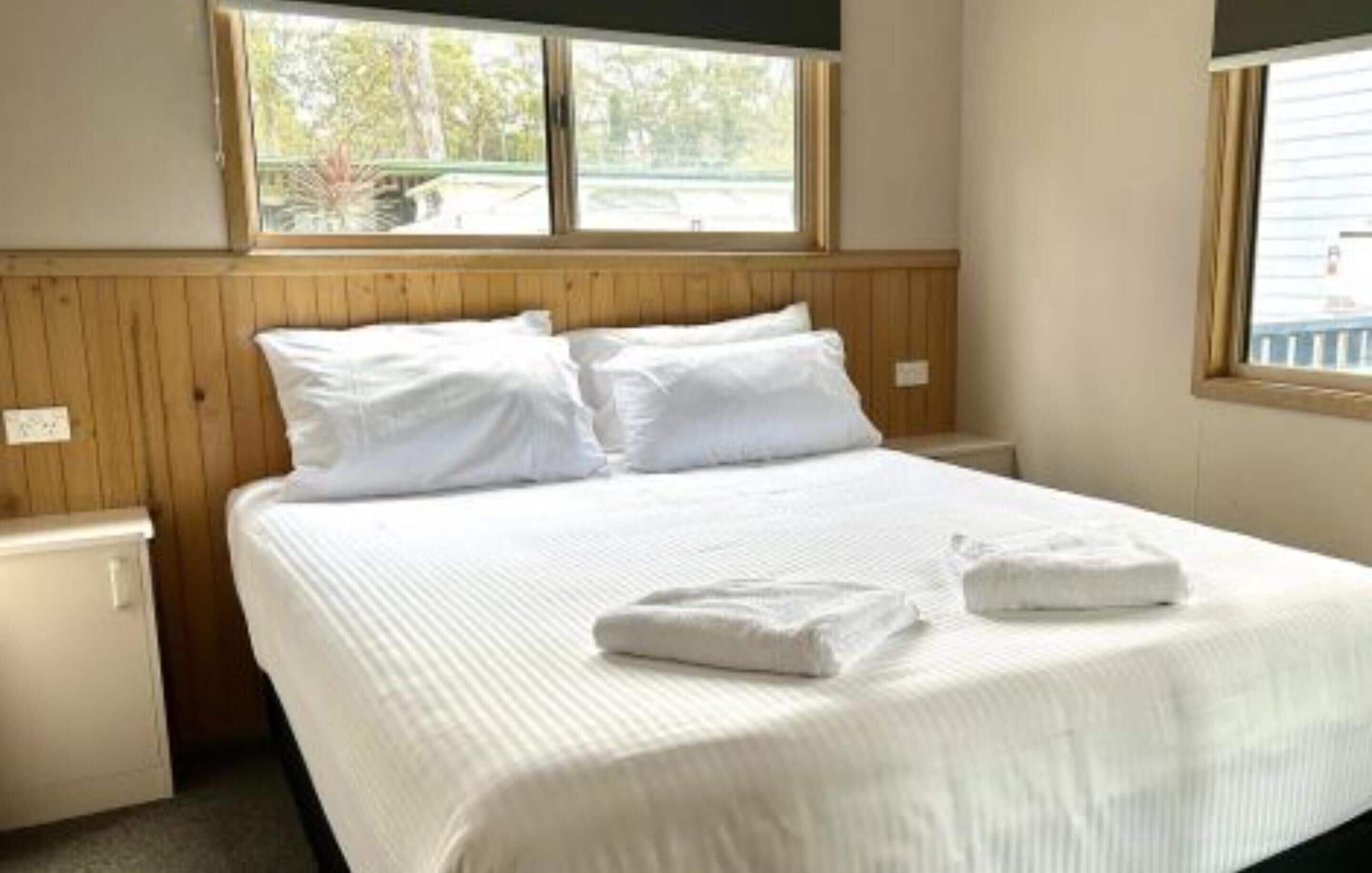 A neatly made bed with white linens and pillows, a wooden headboard, and a window with a view of greenery outside.