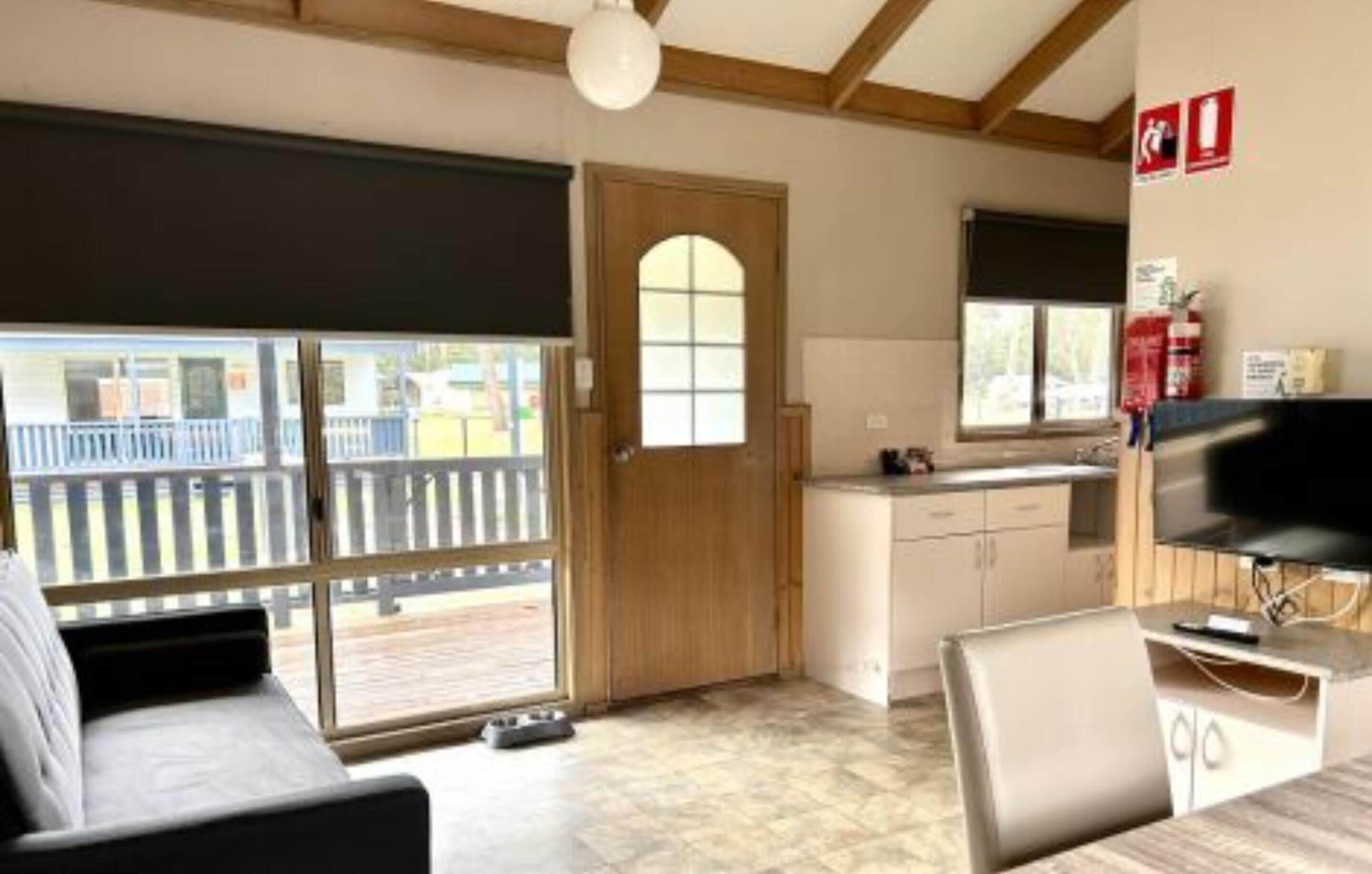 A room with a large sliding glass door leading to a balcony, a kitchenette with a stove and sink, a dining area with a table and chairs, and a television mounted on the wall.