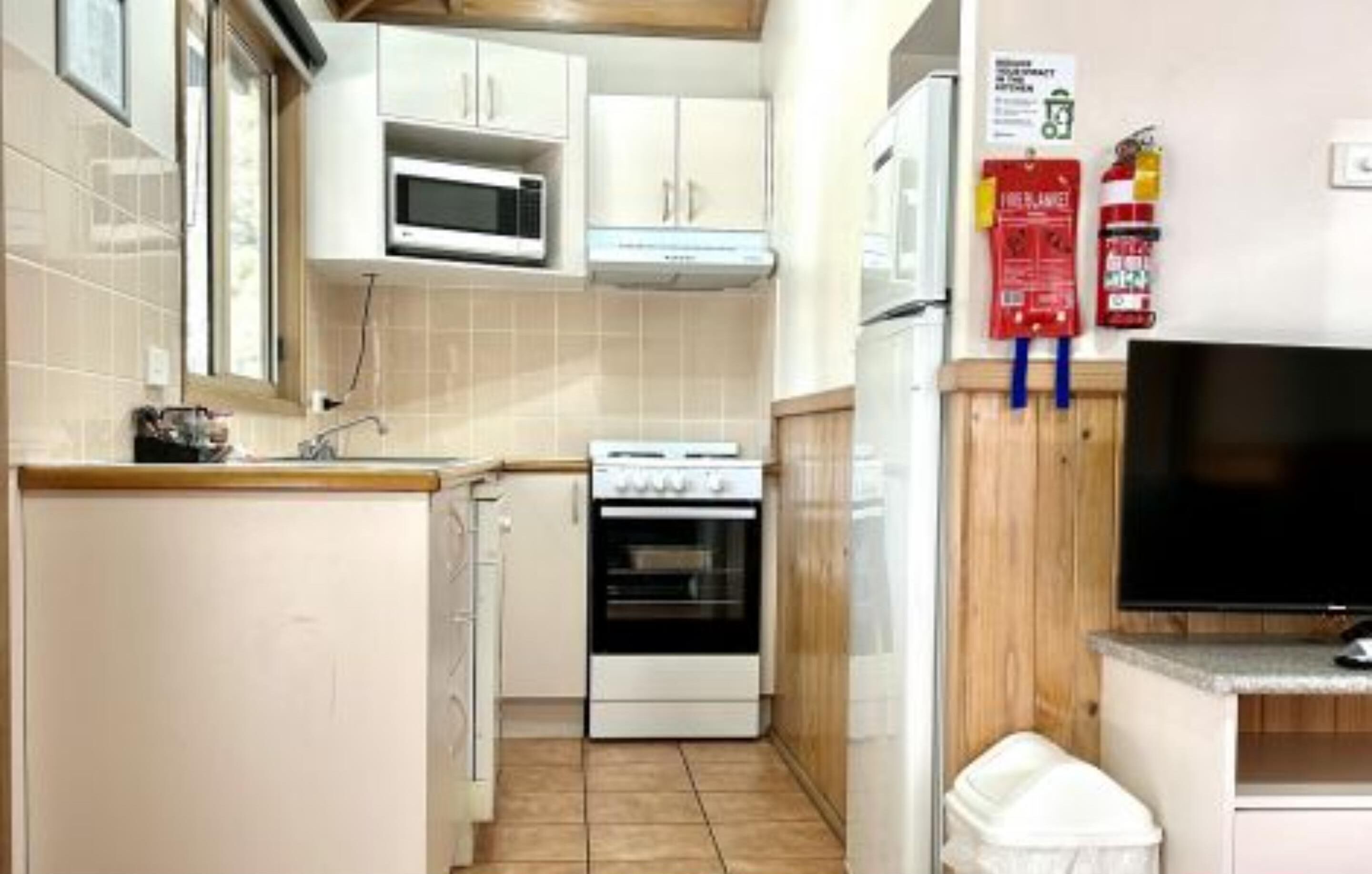 A compact kitchen with white cabinets, a microwave, an oven, and a television on a wooden shelf.
