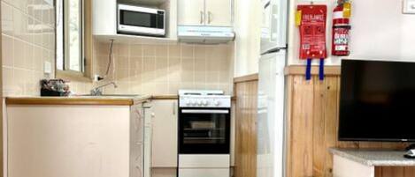 A compact kitchen with white cabinets, a microwave, an oven, and a television on a wooden shelf.