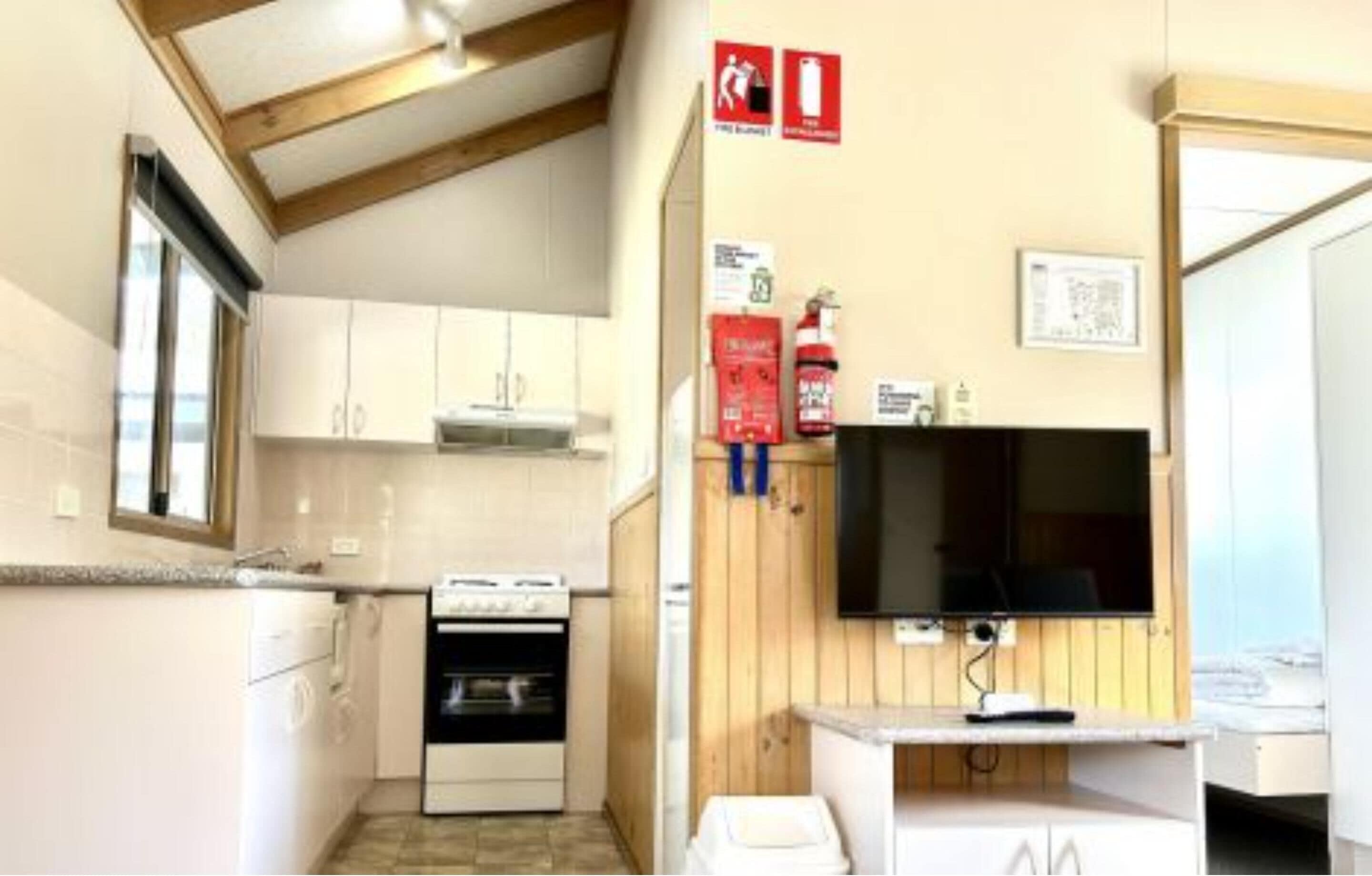 Une cuisine de taille réduite avec des meubles blancs, une cuisinière et un évier. Un mur en bois est orné d’un téléviseur fixé au mur et d’un extincteur.