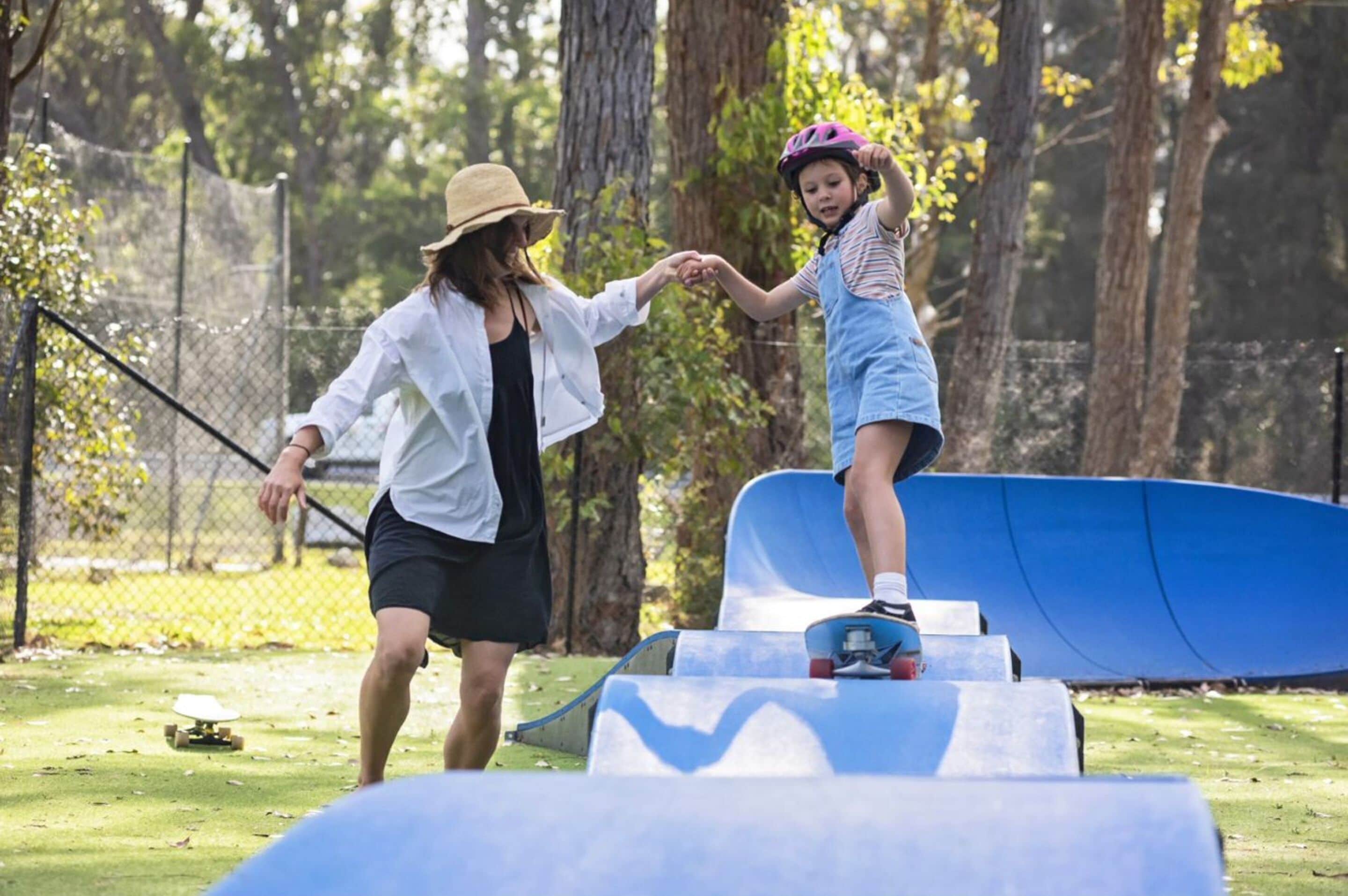 A person in a white shirt and black skirt holding hands with a child on a skateboard ramp in a park.