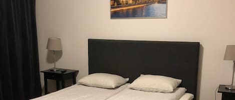 A neatly made bed with a white bedspread and two pillows, a dark headboard, a nightstand with a lamp, and a framed picture of a cityscape on the wall.