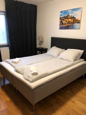 A single bed with a white quilt and two pillows, a bedside table with a lamp, a window with blinds, and a painting of a cityscape on the wall. - Apartment in Årsta Stockholm 238 (Arsta)