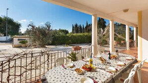 Un área de comedor al aire libre con una mesa preparada para cuatro personas, rodeada de sillas blancas, con vista a un jardín y una calle.