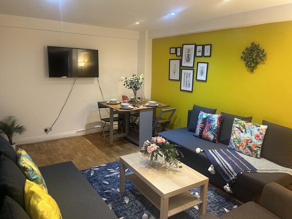 A modern living room with a dining area, a flat-screen TV mounted on the wall, and a vibrant yellow accent wall with framed pictures.