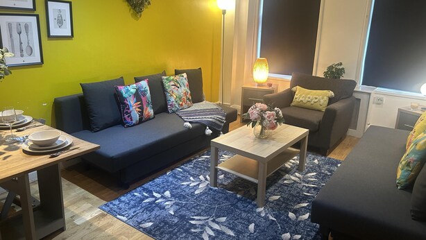 A living room with a blue sofa, a coffee table with a vase of flowers, and a yellow wall with framed pictures. - Marble Arch Family Apartments Posh Central London (London)