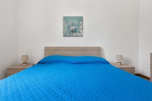 Quarto com colcha azul acolchoada, cabeceira de madeira, duas mesas de cabeceira com luminárias e uma obra de arte na parede acima da cama.