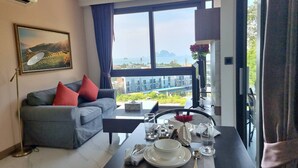 A modern living room with a grey sofa, red cushions, a dining table set with plates, glasses, and cutlery. A large window offers a view of buildings and greenery. - B204 - 1 Bdr Condo With Sea View, Ao Nang Beach (Krabi)