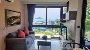 A modern living room with a grey sofa, red cushions, a coffee table, and a large window offering a view of buildings and greenery. - B204 - 1 Bdr Condo With Sea View, Ao Nang Beach (Krabi)