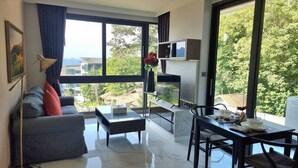 A modern living room with a sofa, coffee table, and dining area, featuring large windows and a view of greenery. - B204 - 1 Bdr Condo With Sea View, Ao Nang Beach (Krabi)