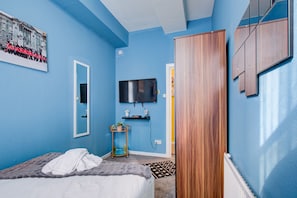 Une chambre à coucher avec un lit, un téléviseur fixé au mur, une armoire en bois et une petite table sur laquelle est posée une plante.