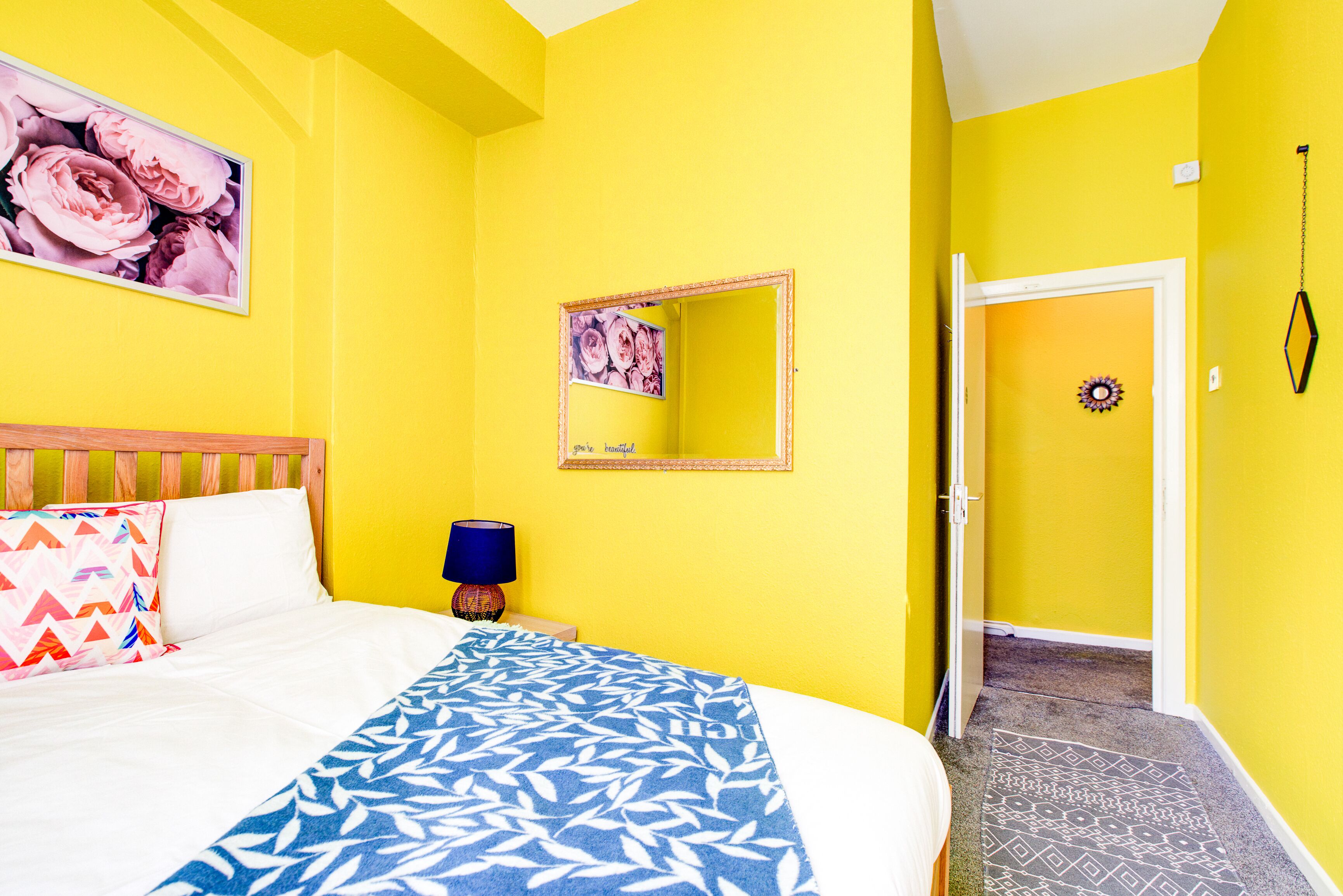 Una stanza luminosa di colore giallo con un letto, uno specchio e una porta che dà su un'altra stanza.