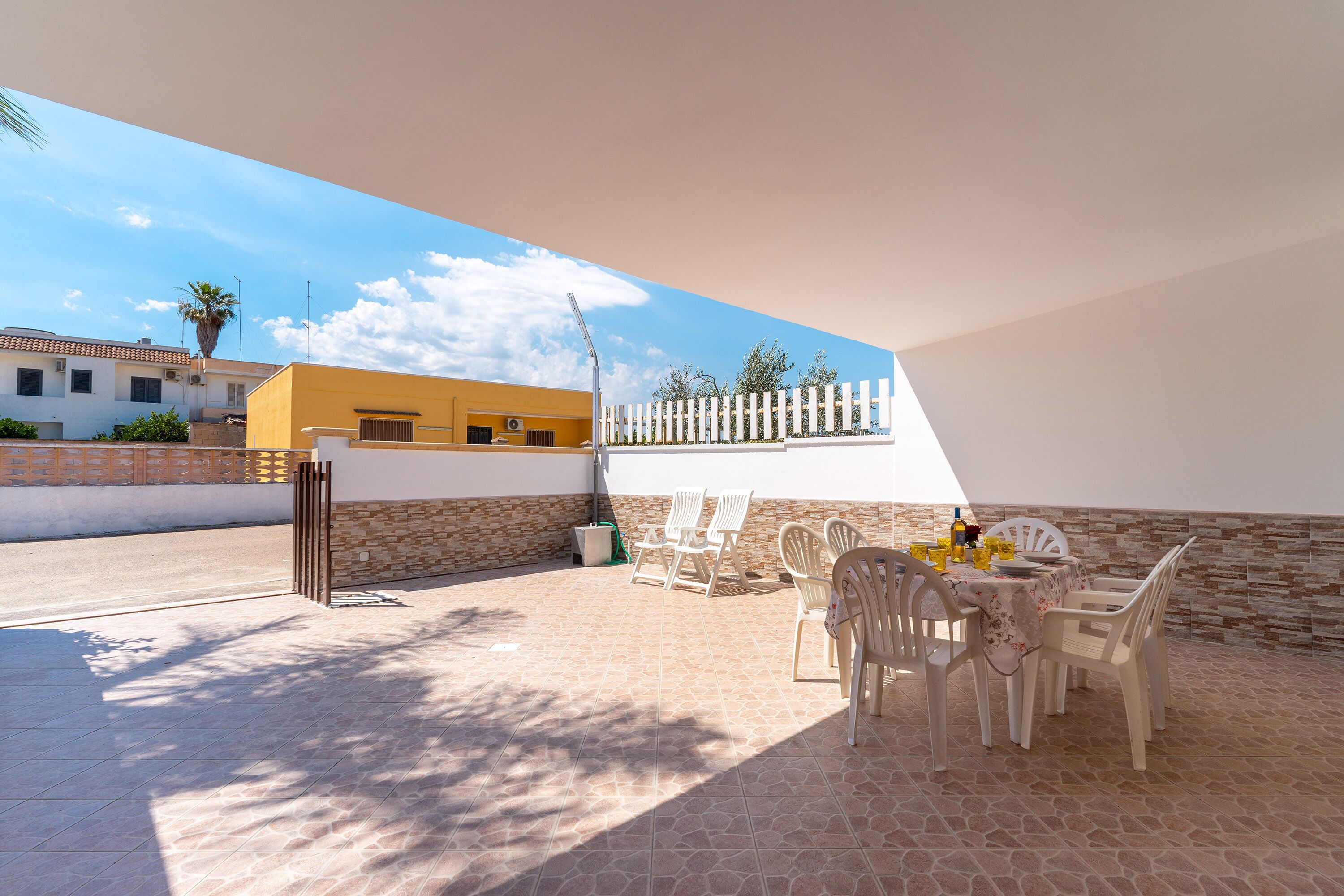 Eine Terrasse im Freien mit Tisch und Stühlen, einem weißen Zaun und einem Gebäude im Hintergrund.